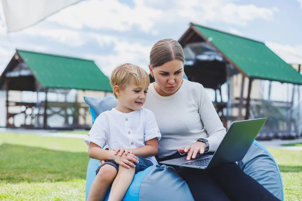 A mother with a laptop, her son sitting next to her outdoors on a green grass lawn. Rest and work with a child