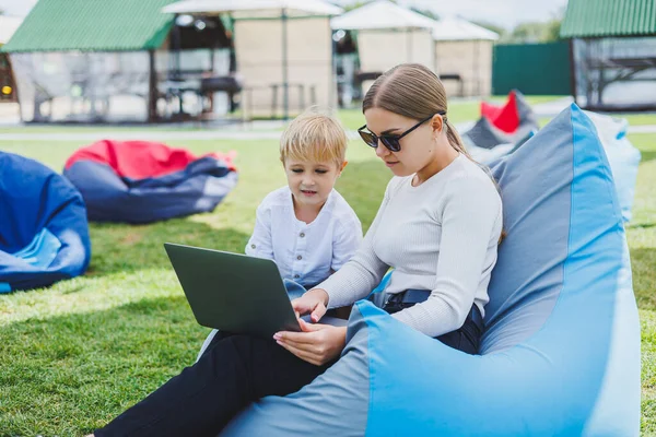 A young woman sits on a soft chair in the park and works on a laptop while her son plays next to her. Working outdoors on maternity leave.