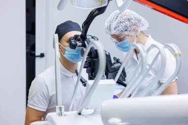 Bir dişçi dişlerini diş mikroskobu ve aletlerle tedavi eder. Asistanın elinde hava ve su dolu bir şırınga var. Diş hekimliği