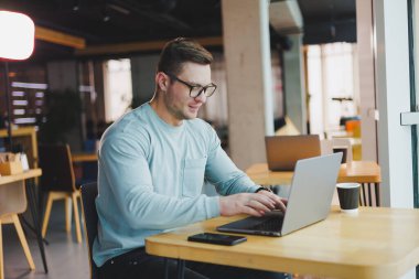 Gündelik giysiler içinde gelecek vaat eden genç bir adam bir masada oturuyor ve bir dizüstü bilgisayar üzerinde çalışıyor. Uzak çalışma için çalışma alanı.