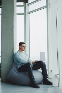 Genç bir yönetici, gözlüklü ve günlük giysileri olan bir adam yumuşak bir sandalyeye oturur ve bir dizüstü bilgisayarda çalışır. Uzak çalışma için çalışma alanı.