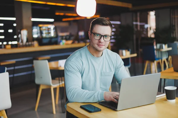A young promising man in casual clothes sits at a table and works on a laptop. Work space for remote work.
