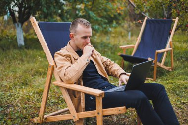 Kahverengi gömlekli yakışıklı bir adam bahçede ahşap bir katlanır sandalyeye oturur ve bir dizüstü bilgisayarla çalışır. Tahta ekolojik mobilyalar. Uzak çalışma
