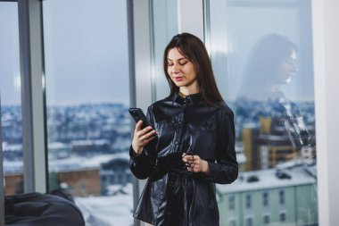 Gündelik giysiler içinde genç zeki bir iş kadını pencerenin yanında durup notlar yazarken ve bulanık modern ofis içlerine karşı çalışırken telefonla çalışıyor..