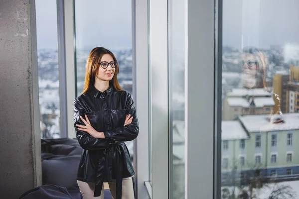 Portrait of a young slender woman in glasses and a black leather shirt. Modern woman on the background of the window in the office with a large window