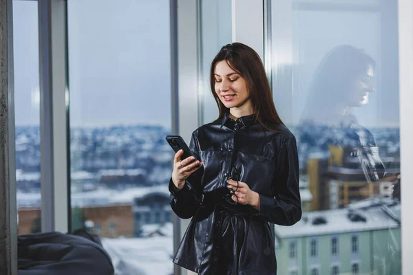 Young smart businesswoman in casual clothes working with phone while standing near window and reporting and writing notes and against blurred modern office interior.