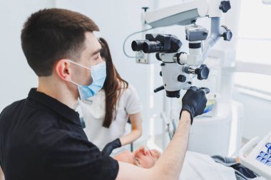 Mikroskoplu modern diş muayenehanesi. Bir dişçi bir adamın dişlerini mikroskop altında tedavi eder, bir asistan ona yardım eder. Diş ağrısı için modern tıbbi bakım