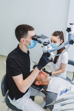 Mikroskoplu modern diş muayenehanesi. Bir dişçi bir adamın dişlerini mikroskop altında tedavi eder, bir asistan ona yardım eder. Diş ağrısı için modern tıbbi bakım