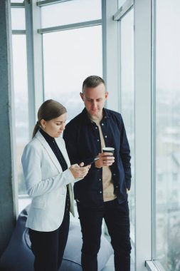 Gülümseyen adam ve kadın, modern şeffaf ofisteki aletlerle pencerenin yanında duruyorlar. Gayri resmi bir ortamda iş arkadaşlarının konuşması.