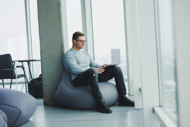 Genç bir yönetici, gözlüklü ve günlük giysileri olan bir adam yumuşak bir sandalyeye oturur ve bir dizüstü bilgisayarda çalışır. Uzak çalışma için çalışma alanı.