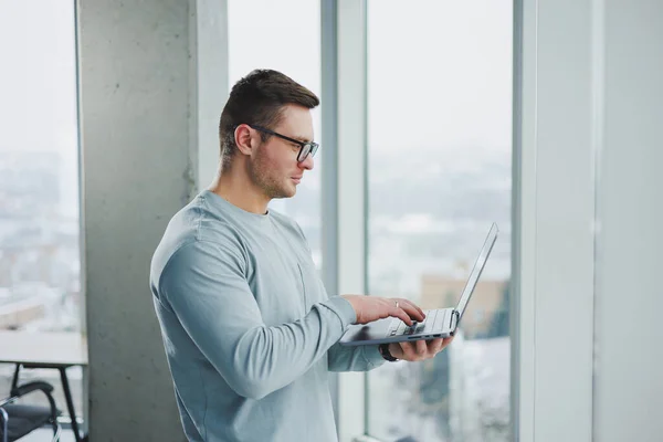 Modern male manager in casual clothes stand by the window in the workspace with a laptop. Male freelancer works remotely.