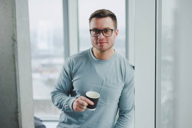 Gündüz vakti modern çalışma alanında pencerenin yanında duran günlük kıyafetlerle gülümseyen adam..