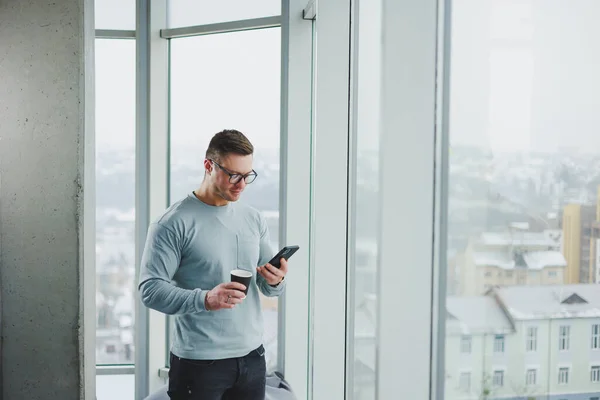 Smiling man in casual clothes standing near window with coffee and looking at smartphone in modern workspace with big window during daytime