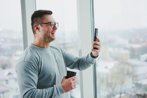 Smiling man in casual clothes standing near window with coffee and looking at smartphone in modern workspace with big window during daytime