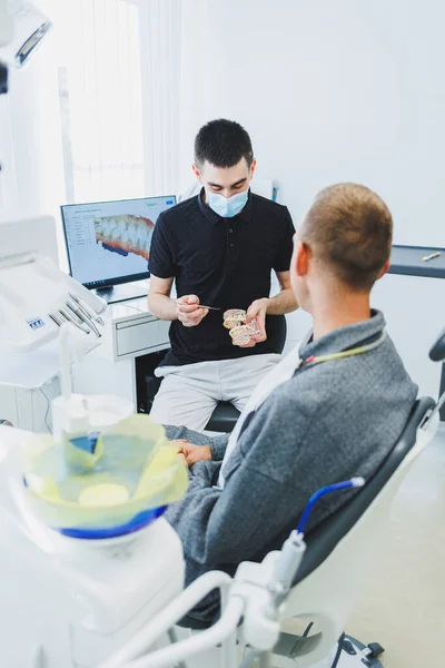 A dentist in a modern dental office tells a patient about dental care. A man sits in a dental chair and listens to a dentist.
