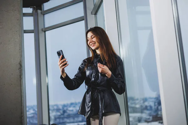 Young smart businesswoman in casual clothes working with phone while standing near window and reporting and writing notes and against blurred modern office interior.