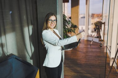 Beyaz ceketli ve gözlüklü, uzun sarı saçlı genç bir iş kadını elinde dizüstü bilgisayarla bir ofis odasında duruyor. Geniş pencereli modern bir ofiste çalış..