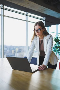 Günlük kıyafetler ve gözlüklerle gülümseyen, beyaz ceket giyen ve çalışırken bir masada dizüstü bilgisayarla gülen genç bir kadın. Ofiste çalış.
