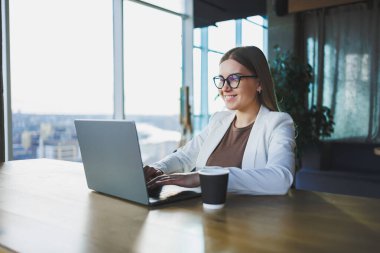 Günlük kıyafetler ve gözlüklerle gülümseyen, beyaz ceket giyen ve çalışırken bir masada dizüstü bilgisayarla gülen genç bir kadın. Ofiste çalış.
