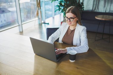 Günlük kıyafetler ve gözlüklerle gülümseyen, beyaz ceket giyen ve çalışırken bir masada dizüstü bilgisayarla gülen genç bir kadın. Ofiste çalış.