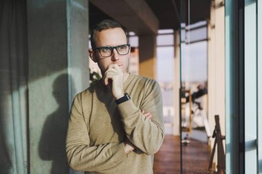 Gözlüklü ve sıradan giyinmiş genç bir adam modern bir çalışma alanında duruyor. Erkek serbest çalışan biri uzaktan çalışıyor..