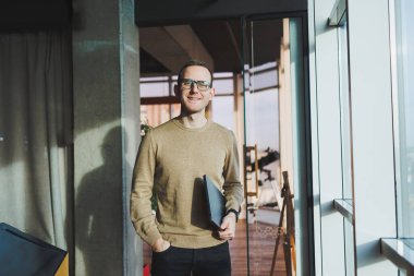 Kahverengi kazaklı ve gözlüklü yakışıklı bir genç ofiste çalışırken elinde dizüstü bilgisayarla büyük bir pencerenin yanında duruyor. Genç bir serbest çalışan uzaktan çalışıyor..