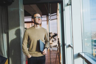 Kahverengi kazaklı ve gözlüklü yakışıklı bir genç ofiste çalışırken elinde dizüstü bilgisayarla büyük bir pencerenin yanında duruyor. Genç bir serbest çalışan uzaktan çalışıyor..