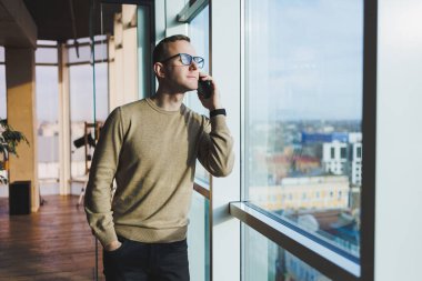 Gözlüklü gülümseyen genç bir adam ofiste dinlenirken telefonla gülümsüyor ve konuşuyor. Genç bir serbest çalışan uzaktan çalışıyor..