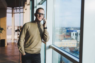 Gözlüklü gülümseyen genç bir adam ofiste dinlenirken telefonla gülümsüyor ve konuşuyor. Genç bir serbest çalışan uzaktan çalışıyor..