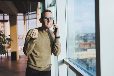 Gözlüklü gülümseyen genç bir adam ofiste dinlenirken telefonla gülümsüyor ve konuşuyor. Genç bir serbest çalışan uzaktan çalışıyor..