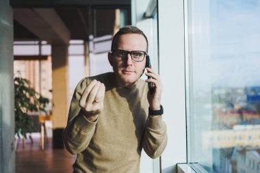 Gözlüklü gülümseyen genç bir adam ofiste dinlenirken telefonla gülümsüyor ve konuşuyor. Genç bir serbest çalışan uzaktan çalışıyor..