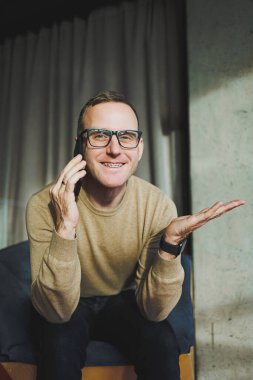 Gözlüklü gülümseyen genç bir adam ofiste dinlenirken telefonla gülümsüyor ve konuşuyor. Genç bir serbest çalışan uzaktan çalışıyor..