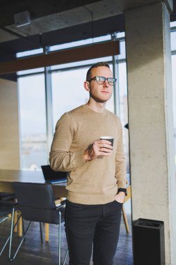 Gözlüklü ve şık giyimli bir işadamı modern bir çalışma alanında kahve içer. Dizüstü bilgisayarı olan serbest çalışan bir erkek.