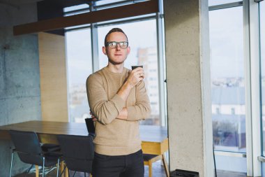 Gözlüklü ve şık giyimli bir işadamı modern bir çalışma alanında kahve içer. Dizüstü bilgisayarı olan serbest çalışan bir erkek.