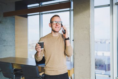 Gözlüklü ve şık giyimli bir işadamı modern bir çalışma alanında kahve içer. Dizüstü bilgisayarı olan serbest çalışan bir erkek.