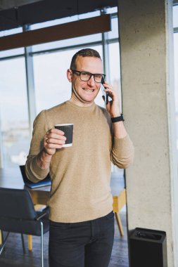 Kahverengi kazaklı ve gözlüklü hoş bir genç adam ofiste dinlenirken duygusal olarak cep telefonuyla konuşuyor ve kahve içiyor. Genç bir serbest çalışan uzaktan çalışıyor..