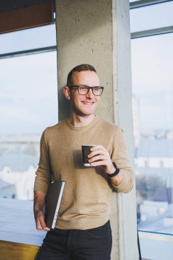 Gözlüklü ve şık giyimli bir işadamı modern bir çalışma alanında kahve içer. Dizüstü bilgisayarı olan serbest çalışan bir erkek.