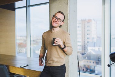 Gözlüklü ve şık giyimli bir işadamı modern bir çalışma alanında kahve içer. Dizüstü bilgisayarı olan serbest çalışan bir erkek.