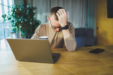 Genç serbest çalışan gözlüklü adam, modern bir çalışma alanında uzaktan kumandalı bir proje üzerinde çalışırken tahta bir masada dizüstü bilgisayar ve kahveyle oturuyor..