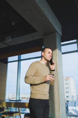 Ofiste dinlenirken gülümseyen, telefonla konuşan ve kahve içen olumlu genç adam. İş sırasında öğle yemeği molası.