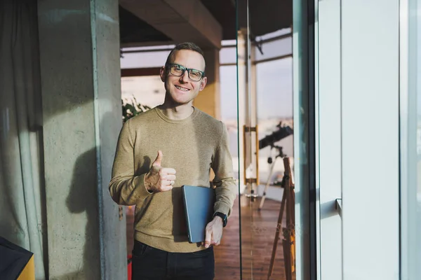 A cute young man in a brown sweater and glasses is standing near a large window in the office with a laptop in his hands while working in the office. A young freelancer works remotely.