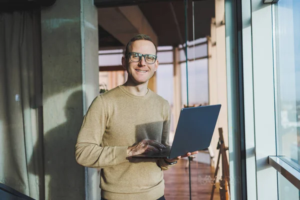 A cute young man in a brown sweater and glasses is standing near a large window in the office with a laptop in his hands while working in the office. A young freelancer works remotely.