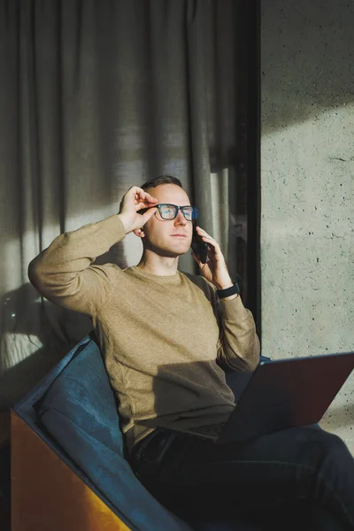 A self-employed man in stylish clothes is making a blog website post on a laptop computer in a modern workspace. Male freelancer works remotely and talks on the phone