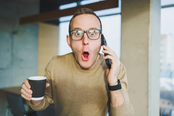 A cute young man in a brown sweater and glasses is talking emotionally on a mobile phone and drinking coffee while relaxing in the office. A young freelancer works remotely.