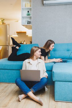Evde çalışan iki genç bayan arkadaş dizüstü bilgisayarların başında kanepede oturuyor. Güzel kız öğrenciler dizüstü bilgisayarlara rapor yazar..