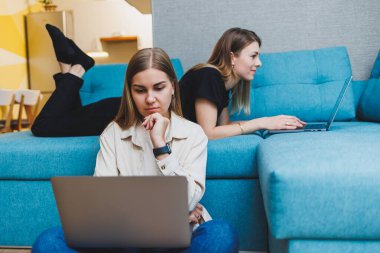 Evde çalışan iki genç bayan arkadaş dizüstü bilgisayarların başında kanepede oturuyor. Güzel kız öğrenciler dizüstü bilgisayarlara rapor yazar..