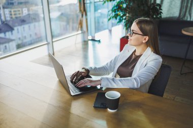 İş kadını, gözlüklü ve günlük kıyafetlerle tahta bir masada dizüstü bilgisayar ve bir fincan kahveyle oturuyor ve dizüstü bilgisayarla ödev yapıyor.
