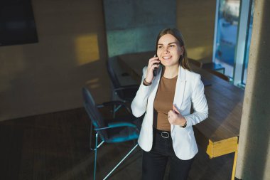 A smiling young woman in a jacket stands in the office and happily talks on the phone. A young manager works in the office and manages the business remotely