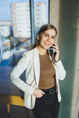 A smiling young woman in a jacket stands in the office and happily talks on the phone. A young manager works in the office and manages the business remotely