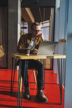 A balanced man in glasses and casual clothes works on a laptop from a cozy workspace. A successful freelancer works remotely.